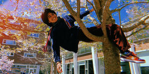 A student sits in the base of a tree with one hand grasping a branch and the other dangling behind her. She smiles at the camera