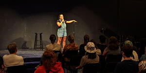 A student stands in the spotlight on stage, performing stand-up comedy for an audience.