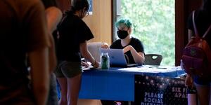 a person sitting behind a laptop wearing a mask speaking to another person across the table