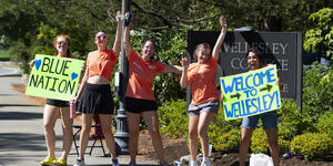 Five students stand at the entrance of Wellesley, waving welcome signs.