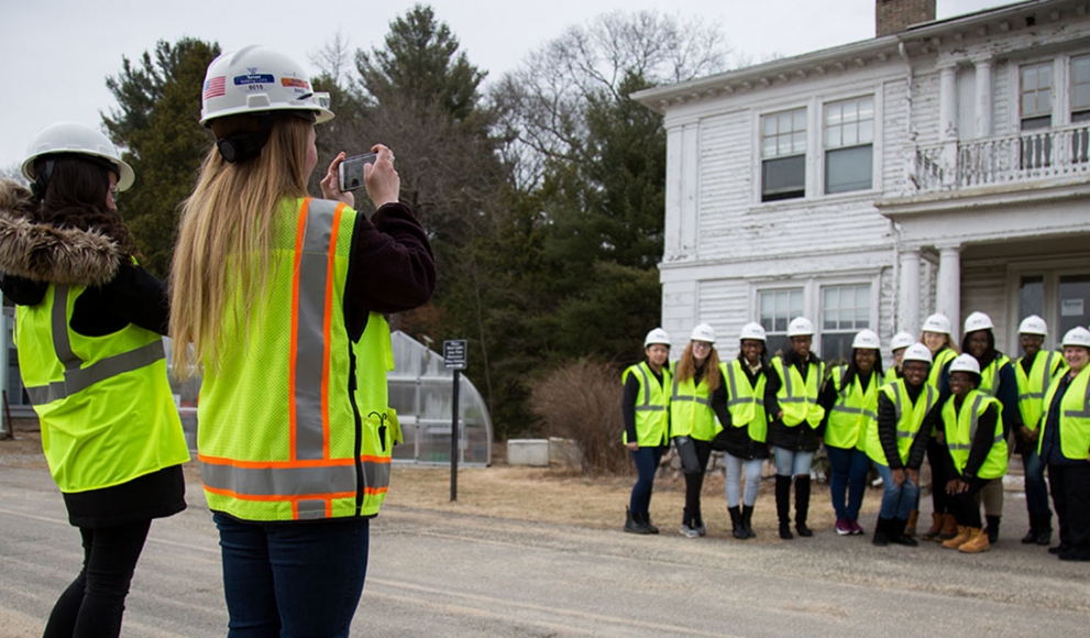A group of students stand outside a building wearing construction vests and hard hats.