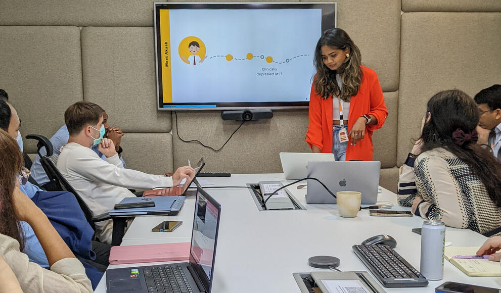 Kavindya Thennakoon leads a meeting. She stands at the front while others sit around a table. A screen is on the wall.