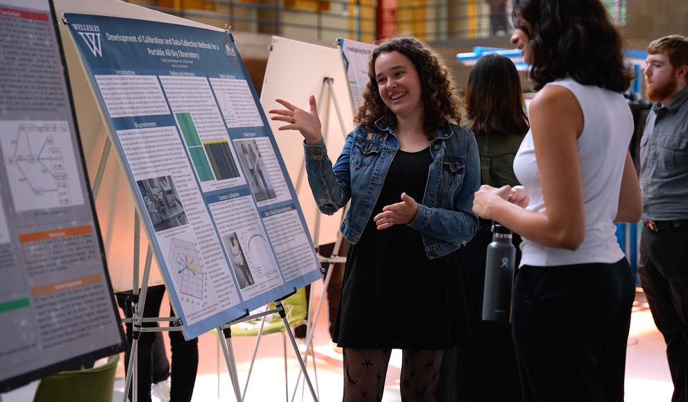 Student gestures toward her science research poster, talking with a visitor.