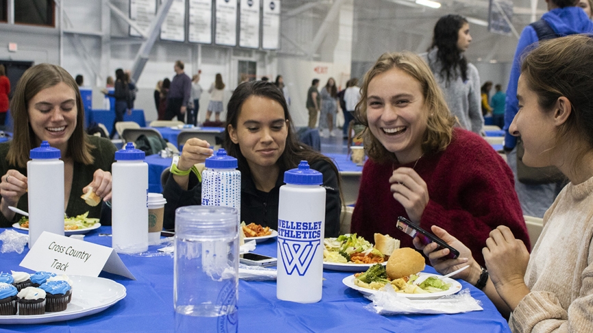 Four students sit at a table in the field house eating pasta