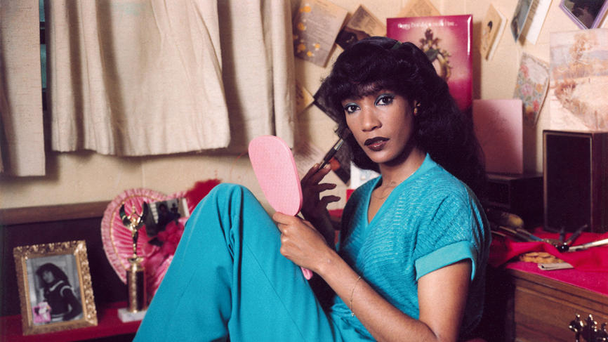 A woman wears a blue jumpsuit and full make up with her legs pulled up under her in a chair. She looks at the camera.