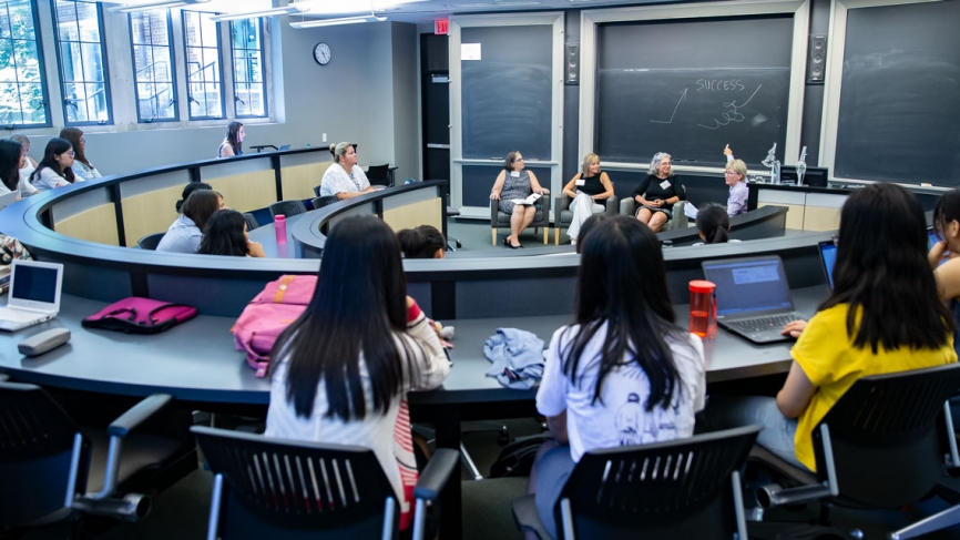 A panel of four women speaks to a crowded classroom.