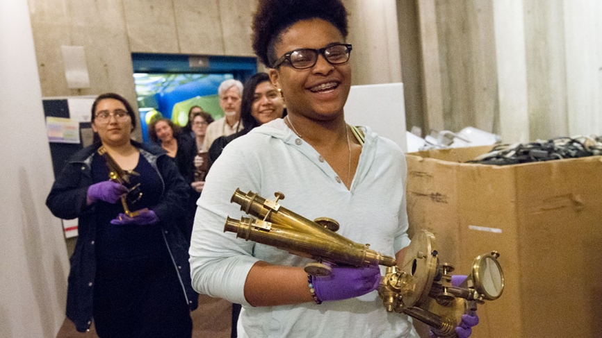 A line of students walk though the basement of the science building wearing purple gloves and holding brass microscopes. 