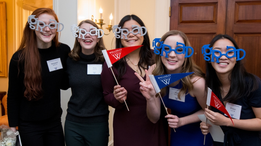 Five students pose for the camera wearing "grad glasses" and holding Wellesley alumnae pennants 
