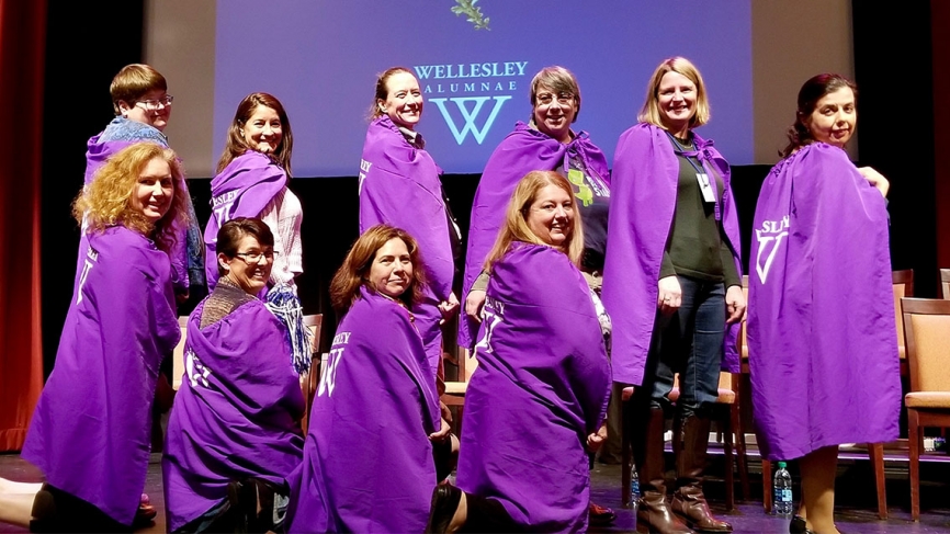 A group of women stand on stage wearing purple capes.