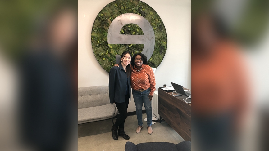 Two women stand in front of a logo that is a backwards "e"