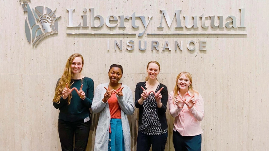 Four women stand in front of a logo that reads "Liberty Mutual." Their hands are in the shape of a W
