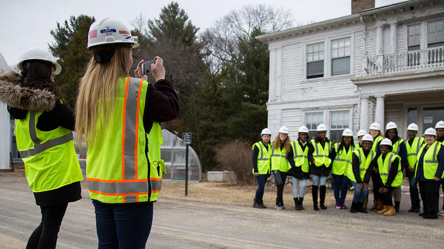 Students stand outside a white building posing in vests and constructions hats.