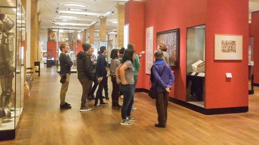 A group of female students stand looking at a painting on a red wall in the German Historical Museum.
