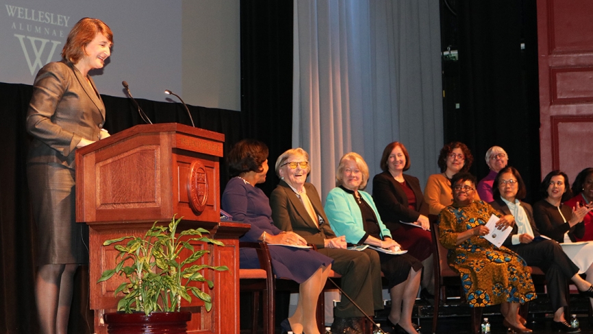 In Alumnae Auditorium, a woman stands at a podium and speaks into a microphone while a group of women sit on stage listening.