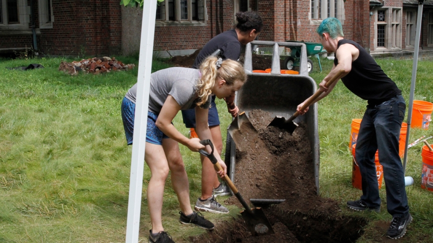 Site workers use shovels to dump dirt from a wheelbarrow into the dig site. 