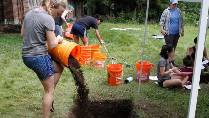 A student pours dirt into a hole from an orange utility bucket. 