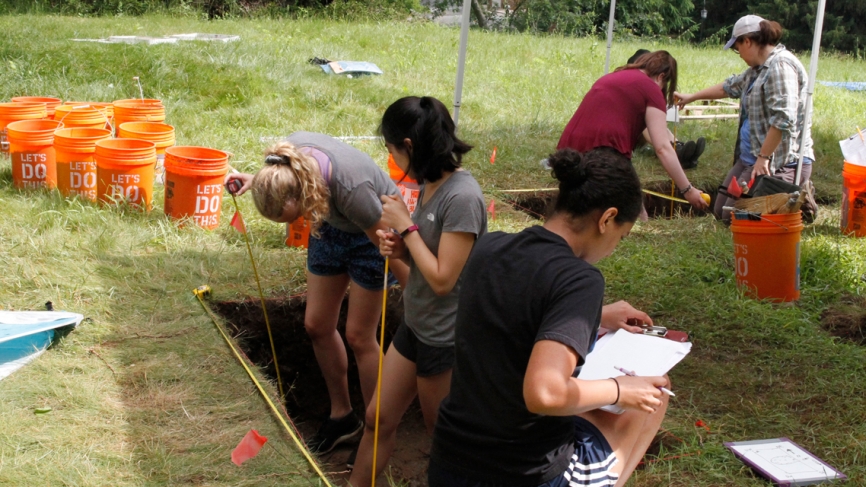 Students measure the dig site and record what they find in white notebooks.