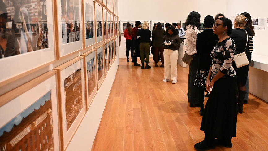 A person stands and looks at a collection of photos hanging on a museum wall. Many people mill around in the background.