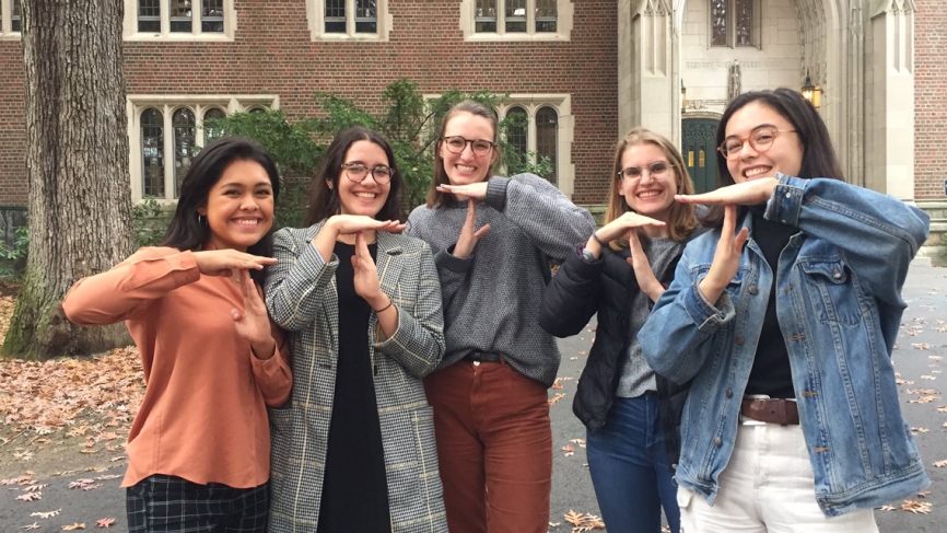 Outside Green Hall, 5 students pose for a photo making a T sign with their hands.