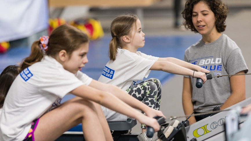 Two young girls sit on ergs as a Wellesley rower looks on