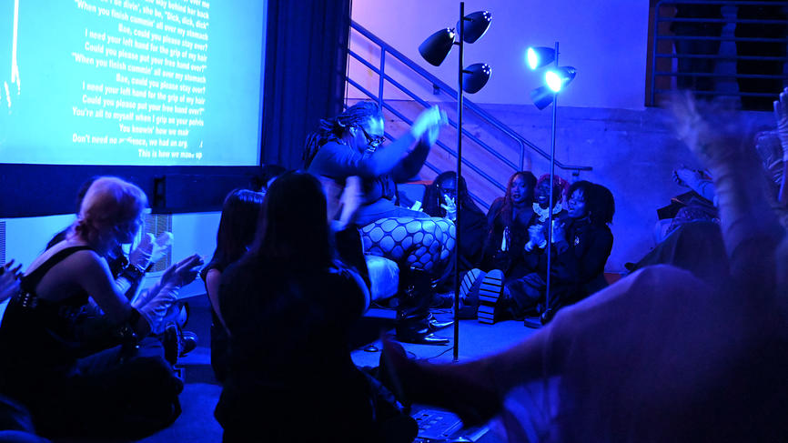 A performance artist is bathed in blue light on stage. She sits in a chair and people sit on the ground around her.