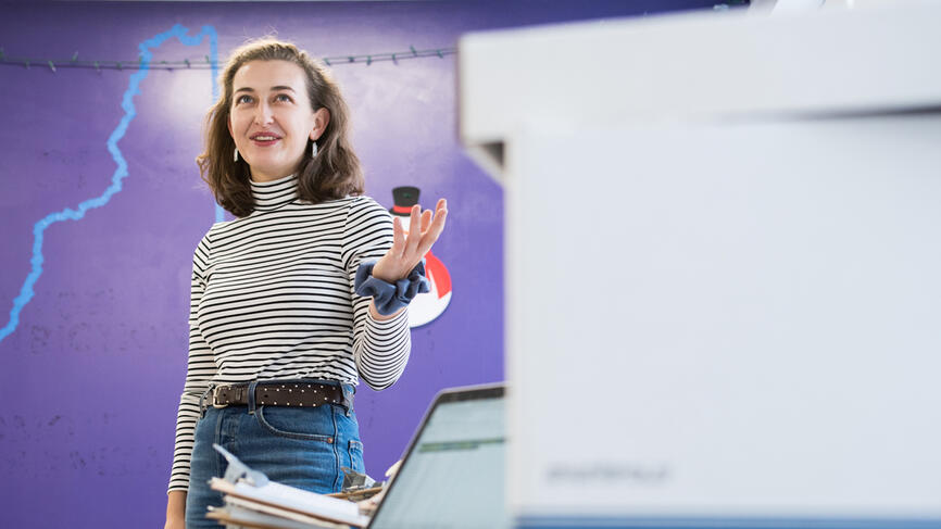 A woman stands in front of a purple wall with the outline of the state of NH in blue tape