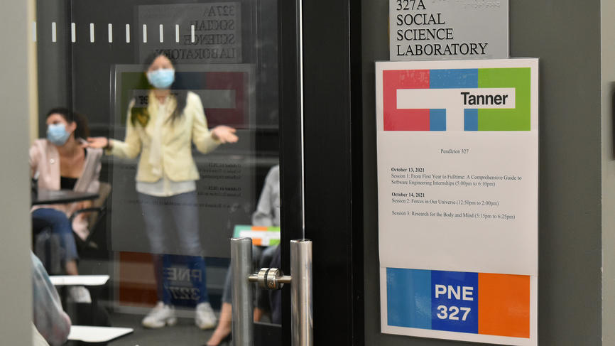 A Tanner promotional sign hangs on the front of a door while in the background a student presents her work.