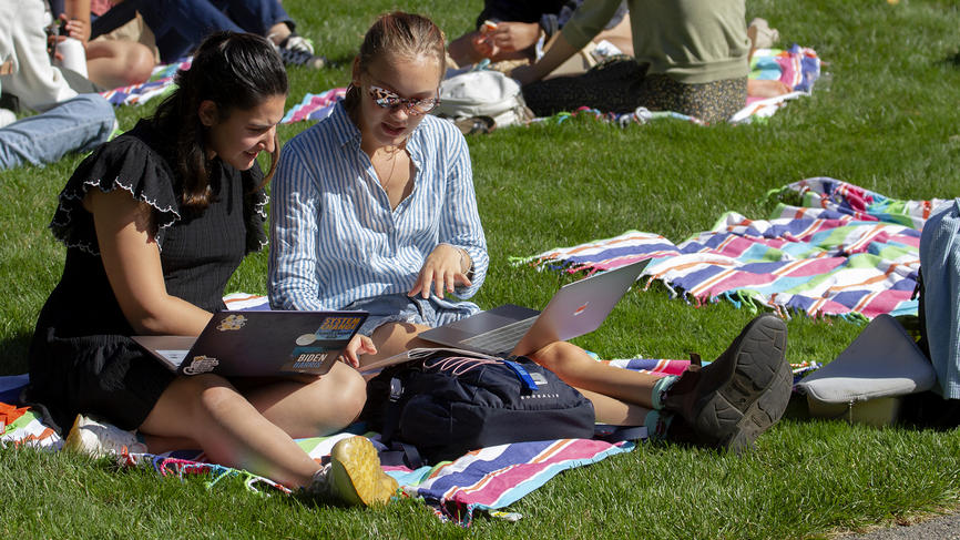 Two students sit on the grass, looking at a computer together.