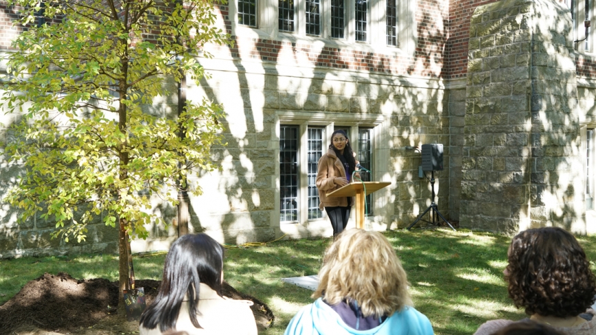 A student speaks at a podium next to a newly planted tree.