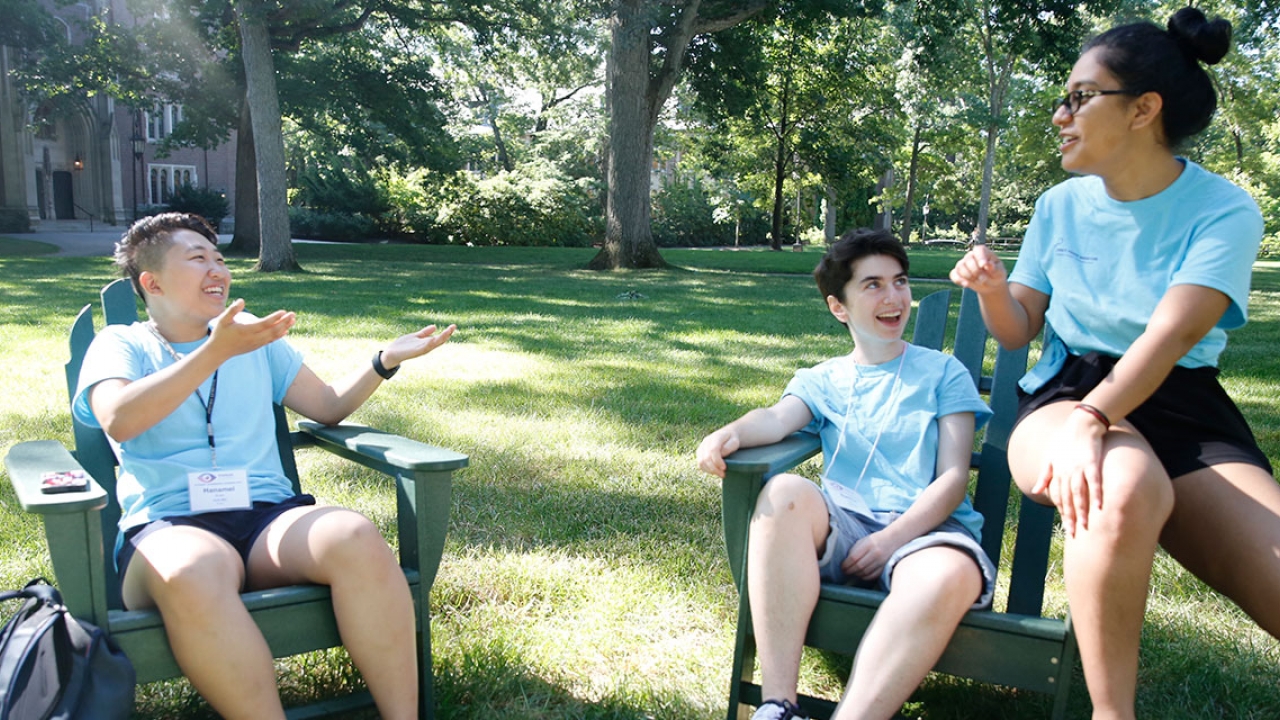 Three students sit at the Academic Quad in Adirondack Chairs, talking.  