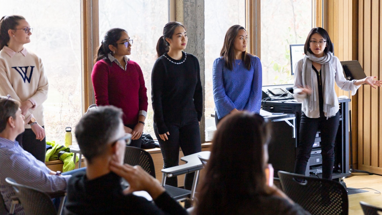 Five students present a project in front of a crowd. One wears a Wellesley "W" sweater.