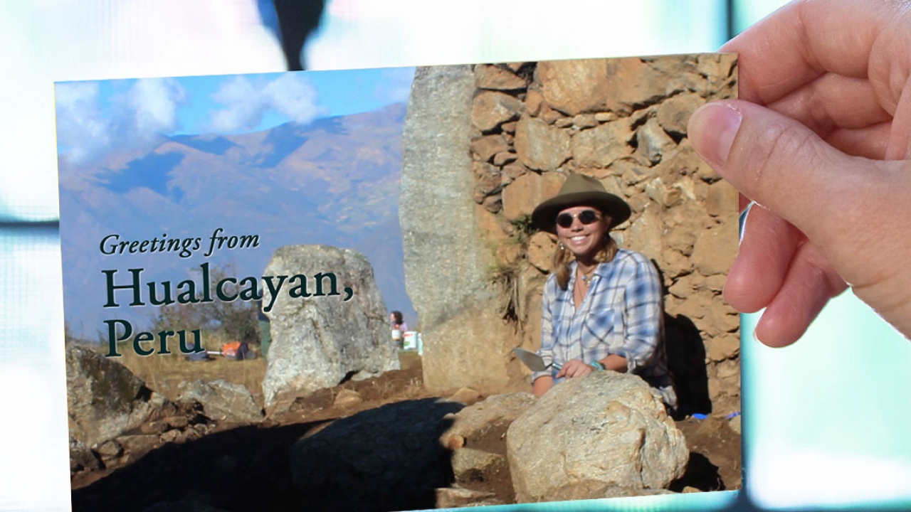 A hand out of frame holds a postcard of a woman sitting at a dig site. It reads, "Greetings from Hualcáyan, Peru."