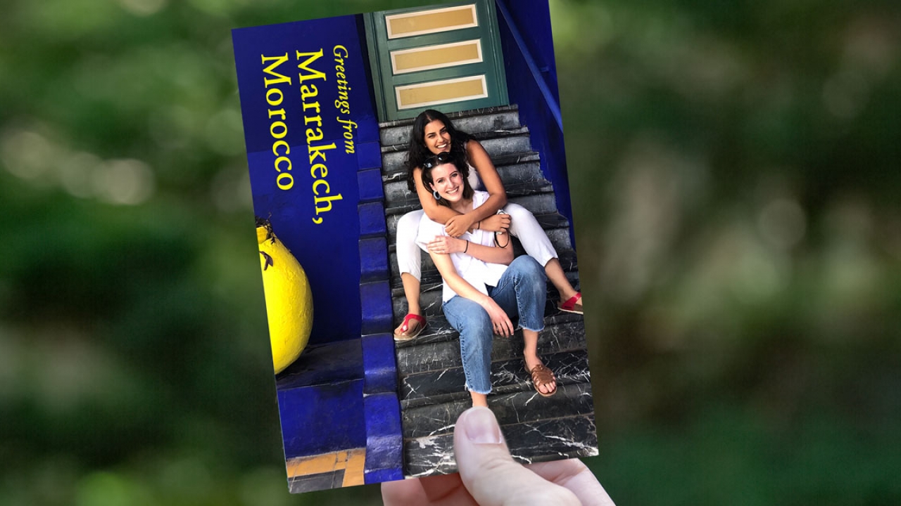 A hand off camera holds a postcard of two students sitting on a staircase. 