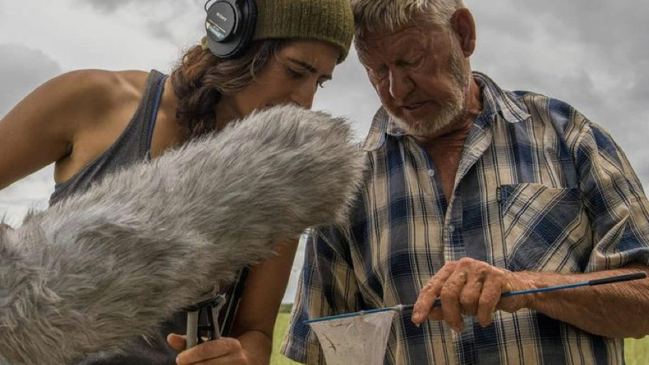 Cat Jaffee holds a microphone and leans toward a man holding a fishing net.