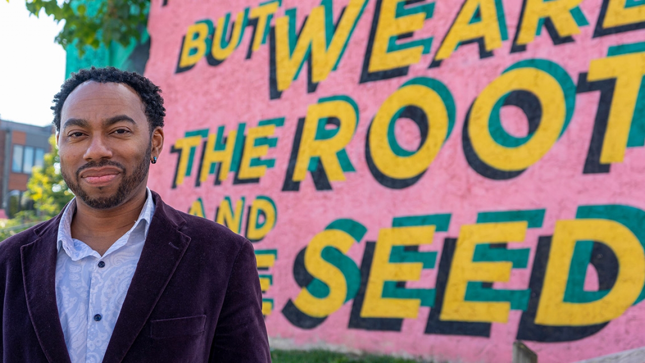 A professor stands in front of public art that reads "we are the root and the seed."