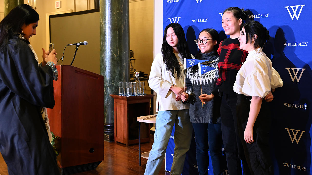 Four December graduates pose for a photo against a Wellesley logo backdrop. 