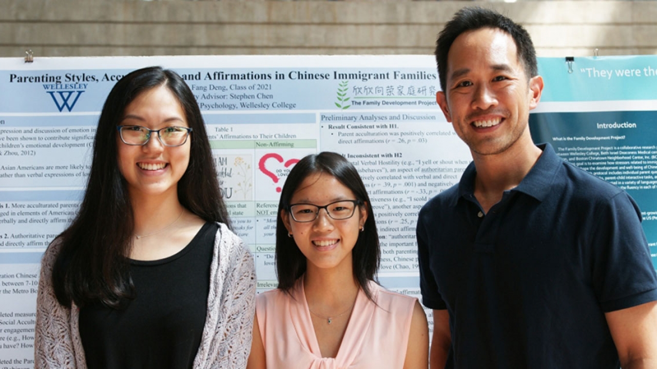 A professor stands in front of a poster with two students. The poster talks about their study.