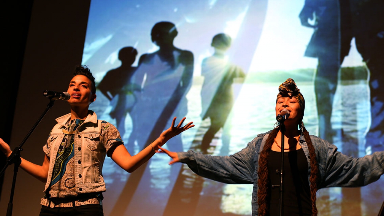 Two performers stand on stage in front of a multimedia presentation.