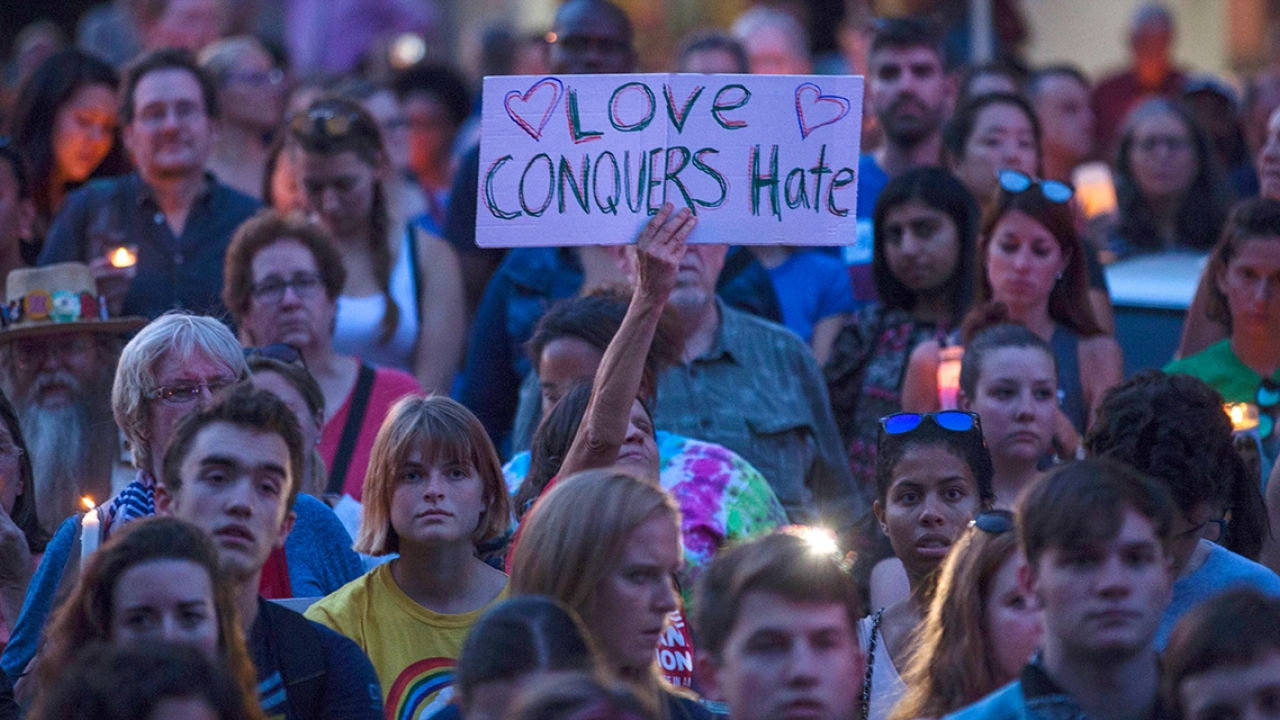 Among a crowd gathered for a vigil, a woman holds a sign that says, "Love Conquers Hate."