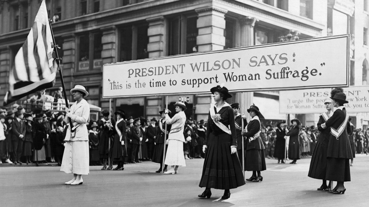 Suffragists march with a sign that reads, "President Wilson says: 'This is the time to support women's suffrage.'