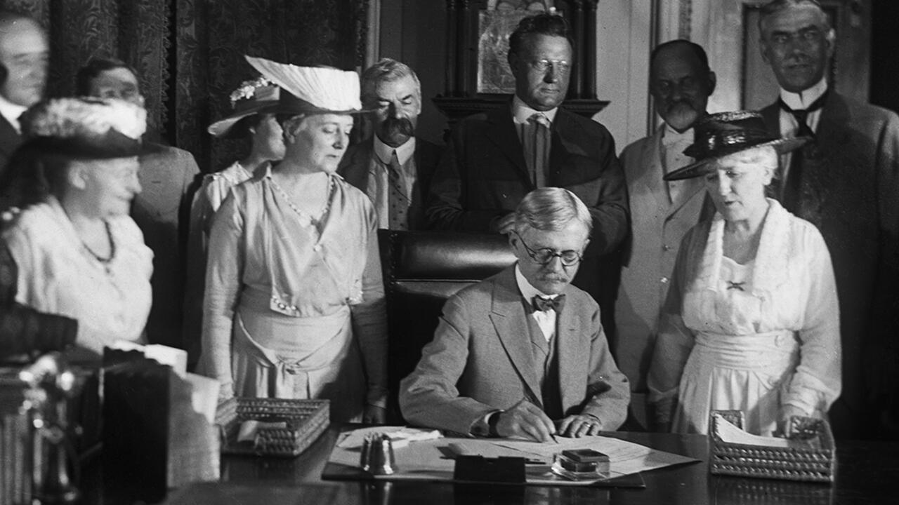 A black and white photo of a man signing a document with women standing behind him. 