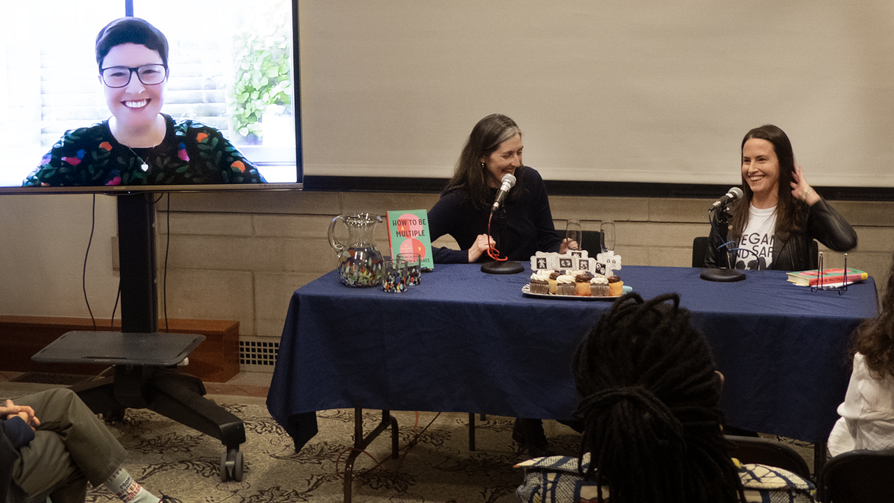 Three speakers at the front of a room including one on a video screen. All three are laughing. 