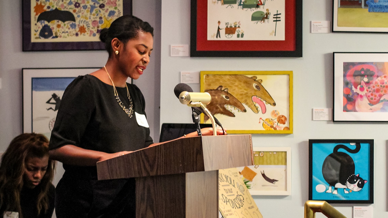 A woman speaks from a podium with artwork all around her