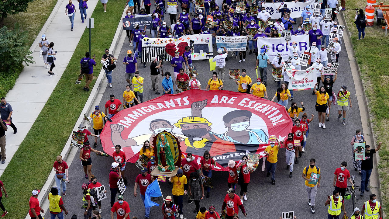 A bird's eye view of a protest march with a participants carrying a sign that says immigrants are essential workers.