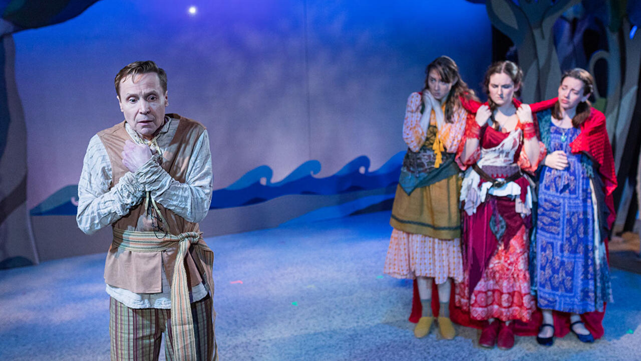 A scene from a play production, three women stand looking at a man.