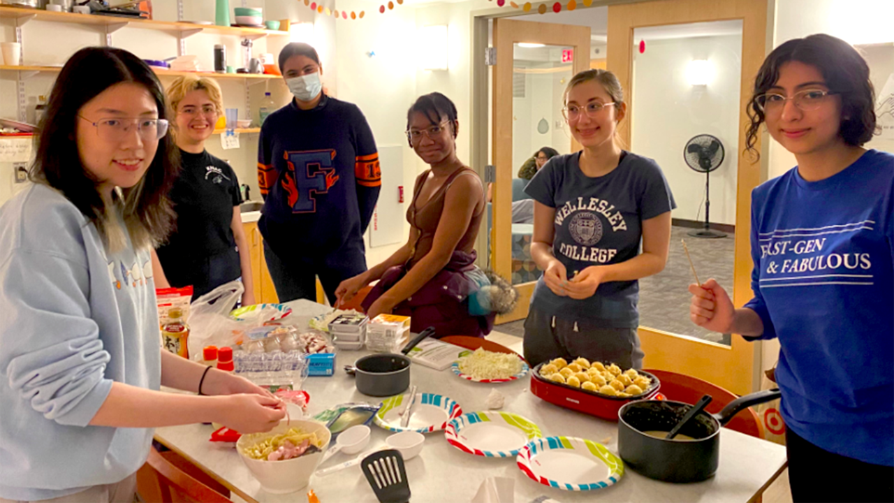 Students gather around a table where food is being made and they are smiling at the camera.