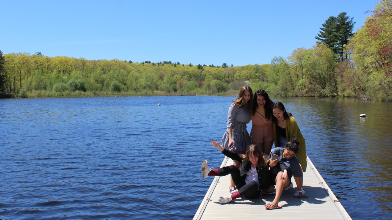 Students stand on a dock by Lake Waban preparing to take a photo. 