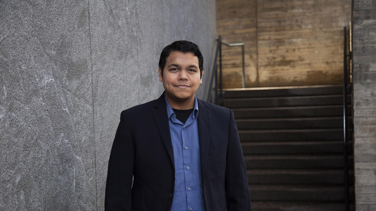 Man in blue shirt and black jacket stands in front of a building.