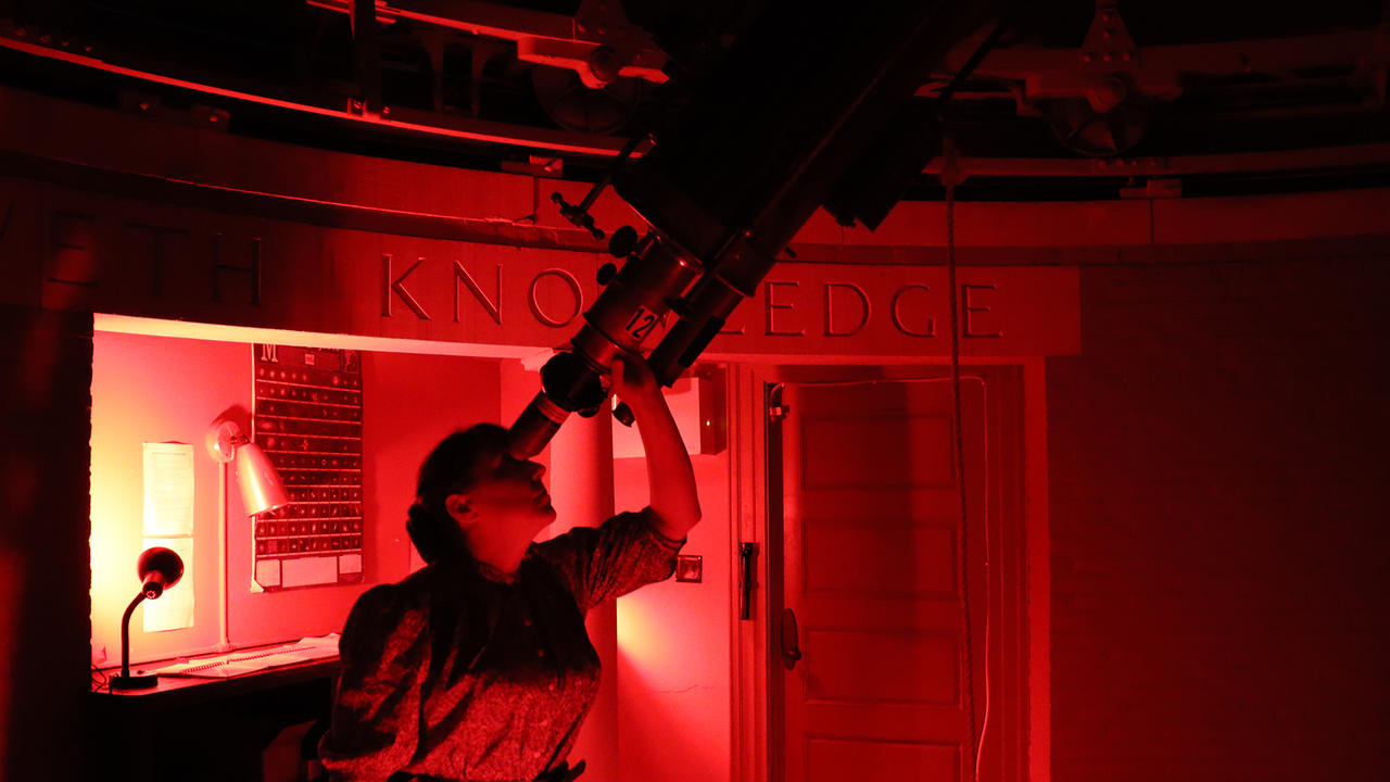 A woman looks through a telescope inside Whitin Observatory. She is bathed in red light. 