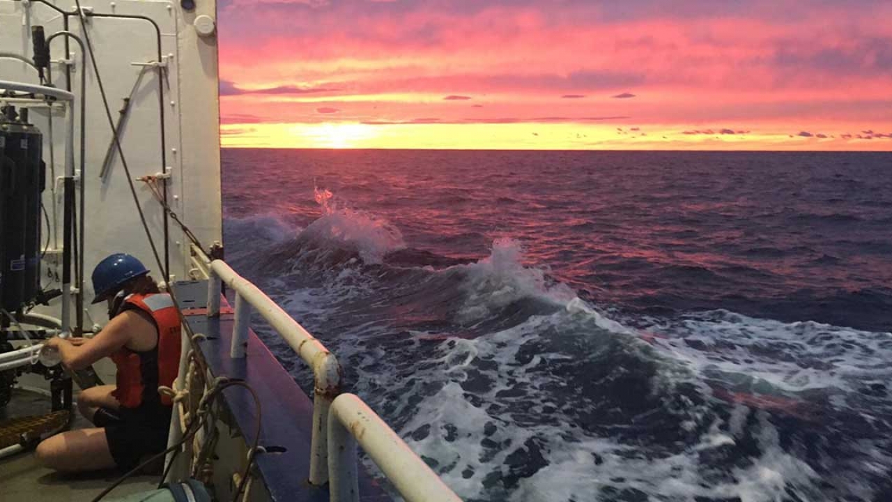 A professor gathers water samples on a boat mid voyage. Photo taken at sunset.